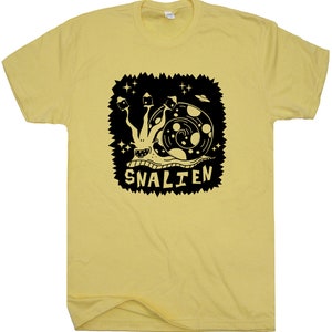 May include: A yellow t-shirt with a black graphic of a snail with a UFO and stars. The text "SNALIEN" is below the snail.