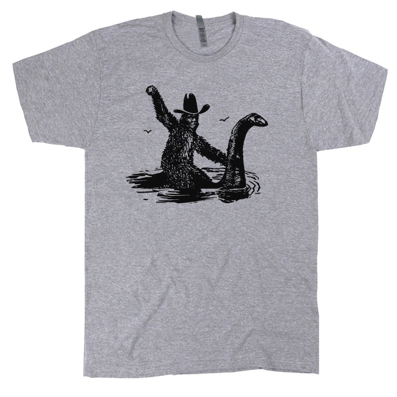 May include: A heather grey t-shirt with a black and white graphic of a creature from the Loch Ness monster riding a cowboy on a lake.