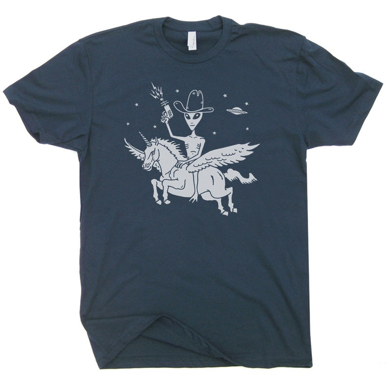 May include: A navy blue t-shirt with a white graphic of an alien wearing a cowboy hat and riding a winged unicorn. The alien is holding a gun and there are stars in the background.