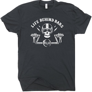 Pu&ograve; includere: T-shirt grigio scuro con grafica bianca. La grafica presenta un teschio con un casco da moto, che tiene un manubrio e la scritta "LIFE BEHIND BARS". Due fulmini su entrambi i lati del teschio.