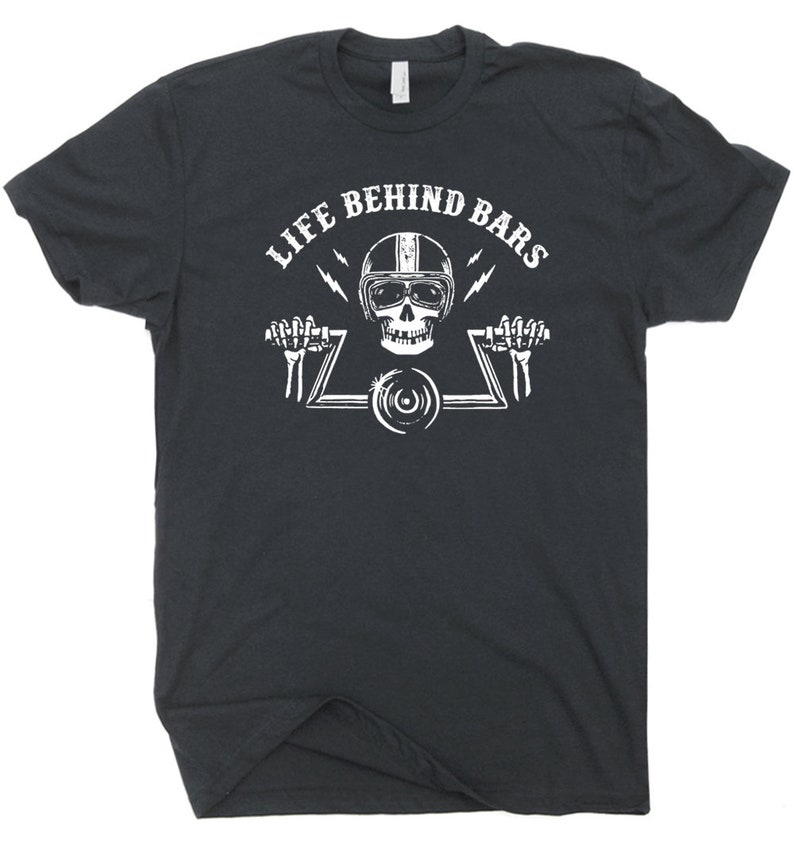 Pu&ograve; includere: T-shirt grigio scuro con grafica bianca. La grafica presenta un teschio con un casco da moto, che tiene un manubrio e la scritta "LIFE BEHIND BARS". Due fulmini su entrambi i lati del teschio.
