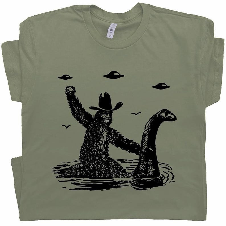 May include: Sage green t-shirt featuring a black graphic of Bigfoot wearing a cowboy hat, embracing the Loch Ness Monster in water, with UFOs in the sky. A humorous design for fans of cryptids and the paranormal.