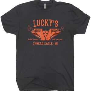 May include: Dark gray t-shirt with an orange graphic. The graphic includes the text "LUCKY'S Custom Cycles" with a stylized design and the words "feelin' lucky... well, are you... SPREAD EAGLE, WI."