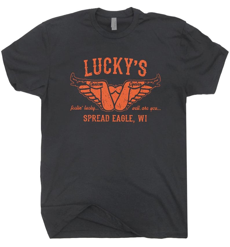 May include: Dark gray t-shirt with an orange graphic. The graphic includes the text "LUCKY'S Custom Cycles" with a stylized design and the words "feelin' lucky... well, are you... SPREAD EAGLE, WI."