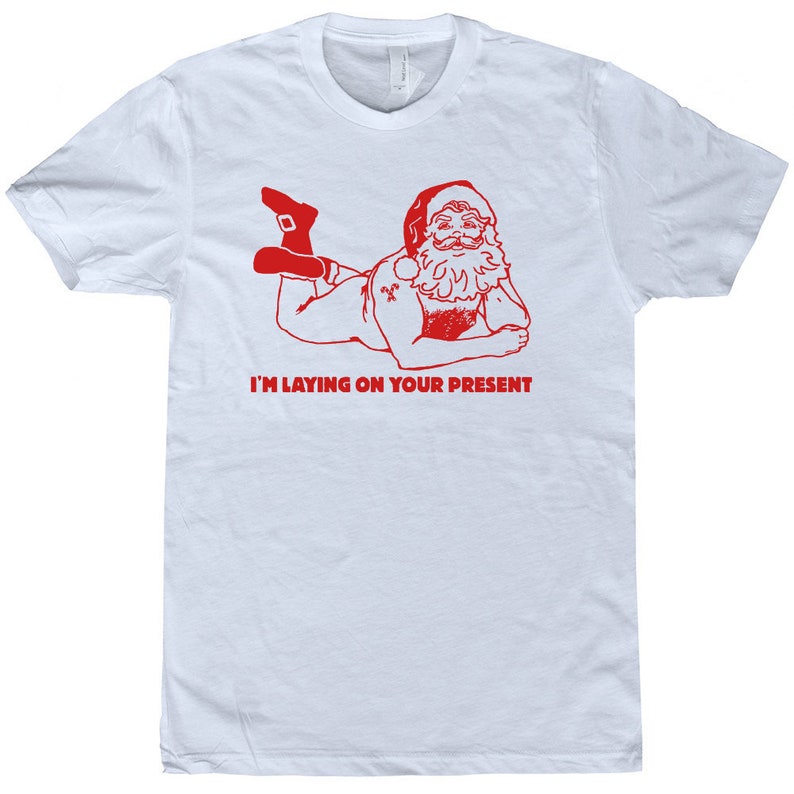 以下が含まれることがあります： 白いTシャツに、サンタクロースがうつ伏せになっている赤いイラストが描かれており、その下に「I'm laying on your present」という文字が書かれています。