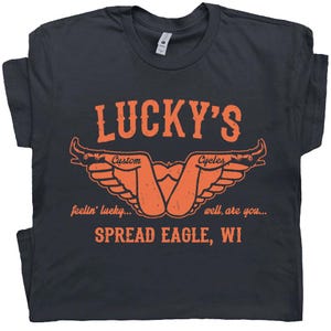 May include: Dark gray t-shirt with an orange graphic. The graphic includes the text "LUCKY'S" above a stylized design of wings and the words "Custom Cycles". Below the design are the words "feelin' lucky... well, are you... SPREAD EAGLE, WI".