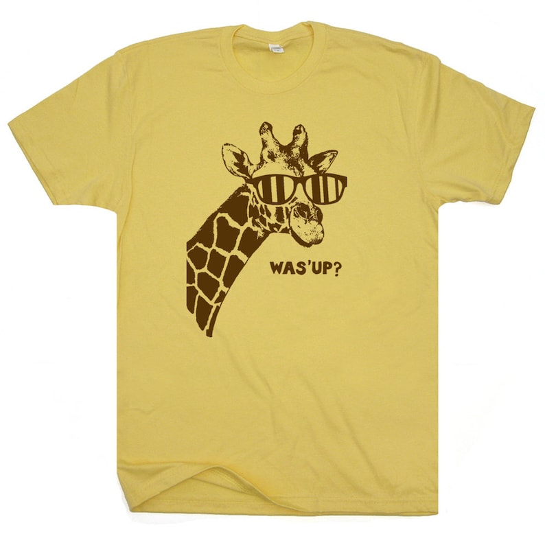 Puede incluir: Una camiseta amarilla con una jirafa marr&oacute;n que lleva gafas de sol y el texto "WAS'UP?" impreso en la parte delantera.