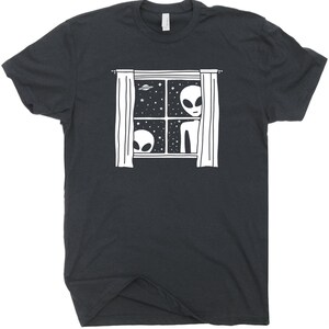 May include: A dark grey t-shirt featuring a white graphic of two aliens peering out of a window with curtains. The window is framed and has stars and a UFO in the background.