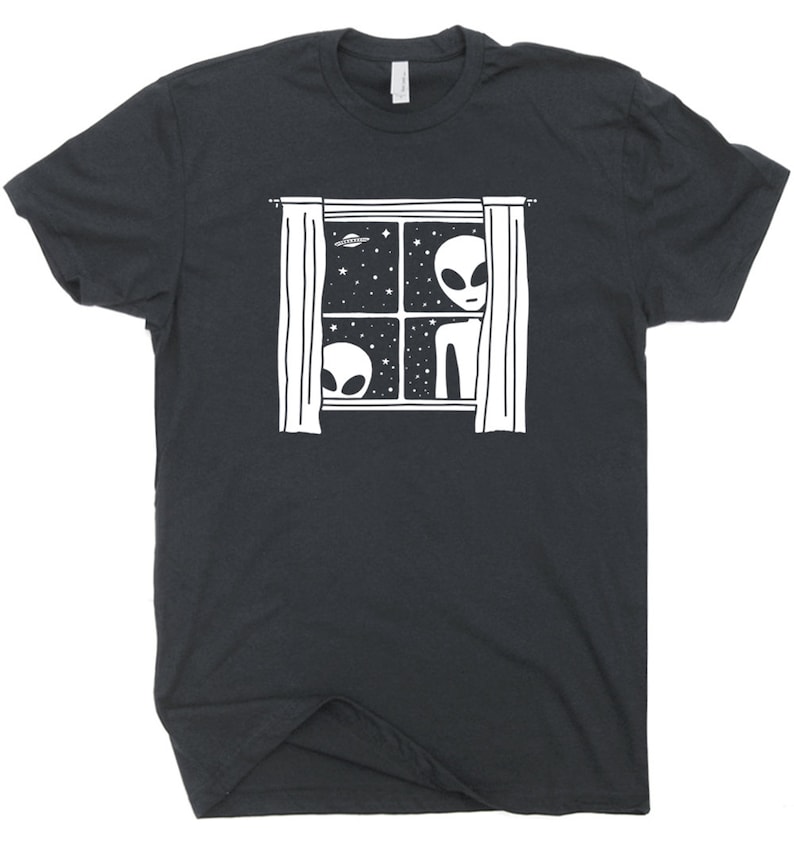 May include: A dark grey t-shirt featuring a white graphic of two aliens peering out of a window with curtains. The window is framed and has stars and a UFO in the background.