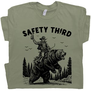 May include: Sage green t-shirt with a black graphic of a cowboy riding a bear, holding a bottle. The text "SAFETY THIRD" is above the graphic, with a lightning bolt replacing the "I". The design includes trees and birds.