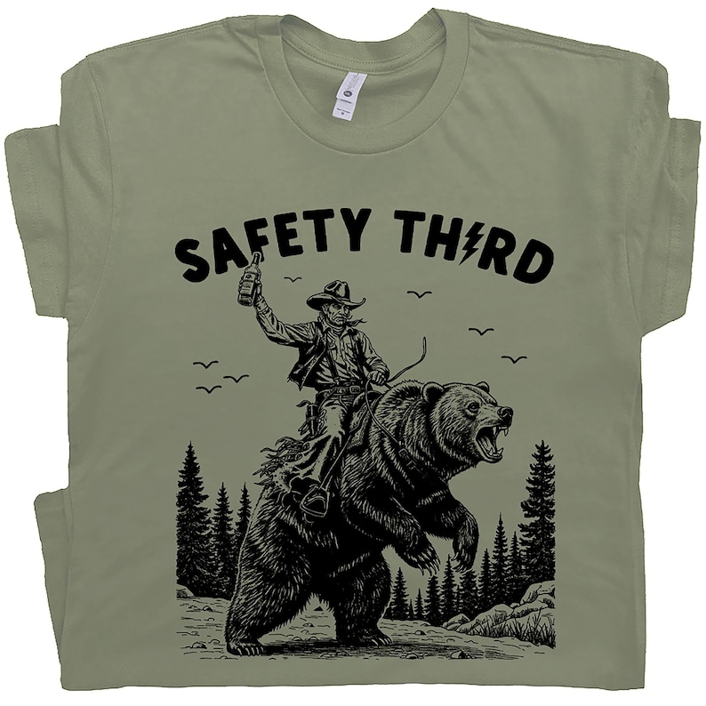 May include: Sage green t-shirt with a black graphic of a cowboy riding a bear, holding a bottle. The text "SAFETY THIRD" is above the graphic, with a lightning bolt replacing the "I". The design includes trees and birds.