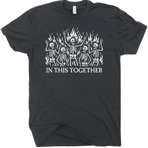 May include: Dark gray t-shirt featuring a white graphic of skeletons in flames. The skeletons are waving, and the text "IN THIS TOGETHER" is printed below the graphic. The shirt has a crew neck and short sleeves.