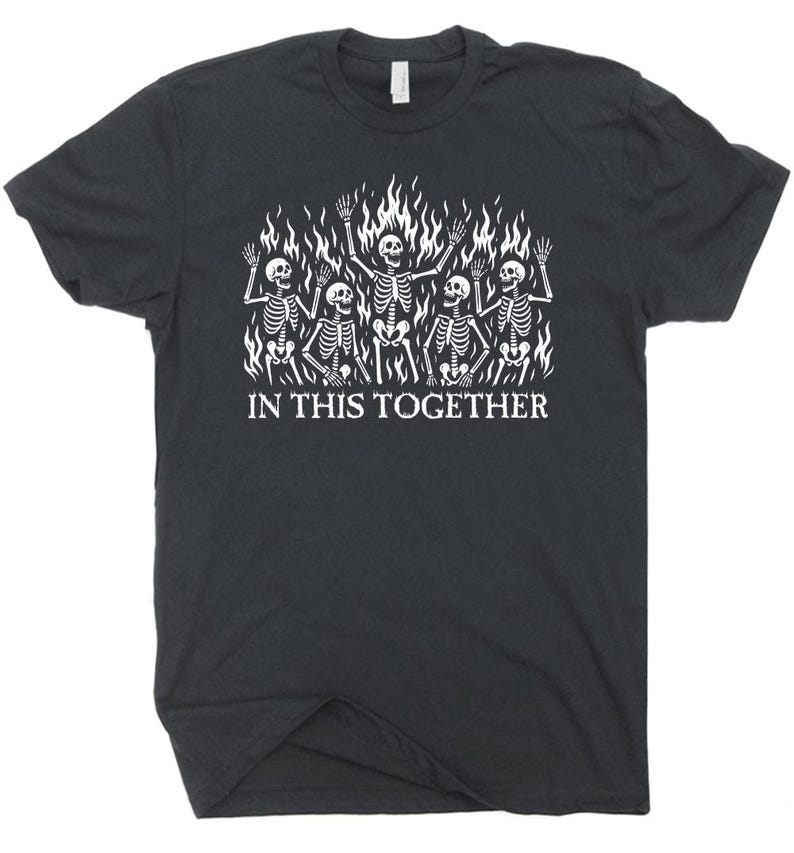 May include: Dark gray t-shirt featuring a white graphic of skeletons in flames. The skeletons are waving, and the text "IN THIS TOGETHER" is printed below the graphic. The shirt has a crew neck and short sleeves.