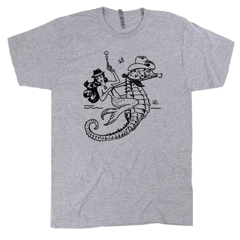Op de afbeelding: Een grijs T-shirt met een zwart-witte illustratie van een zeemeermin die op een zeepaardje rijdt. De zeemeermin draagt een hoed en houdt een pistool vast.