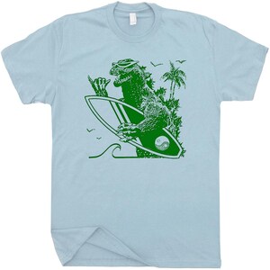 Puede incluir: Camiseta azul claro con un gr&aacute;fico verde de Godzilla con gafas de sol y una tabla de surf. La tabla de surf tiene un s&iacute;mbolo de yin-yang. Tambi&eacute;n se representan palmeras, olas y p&aacute;jaros.