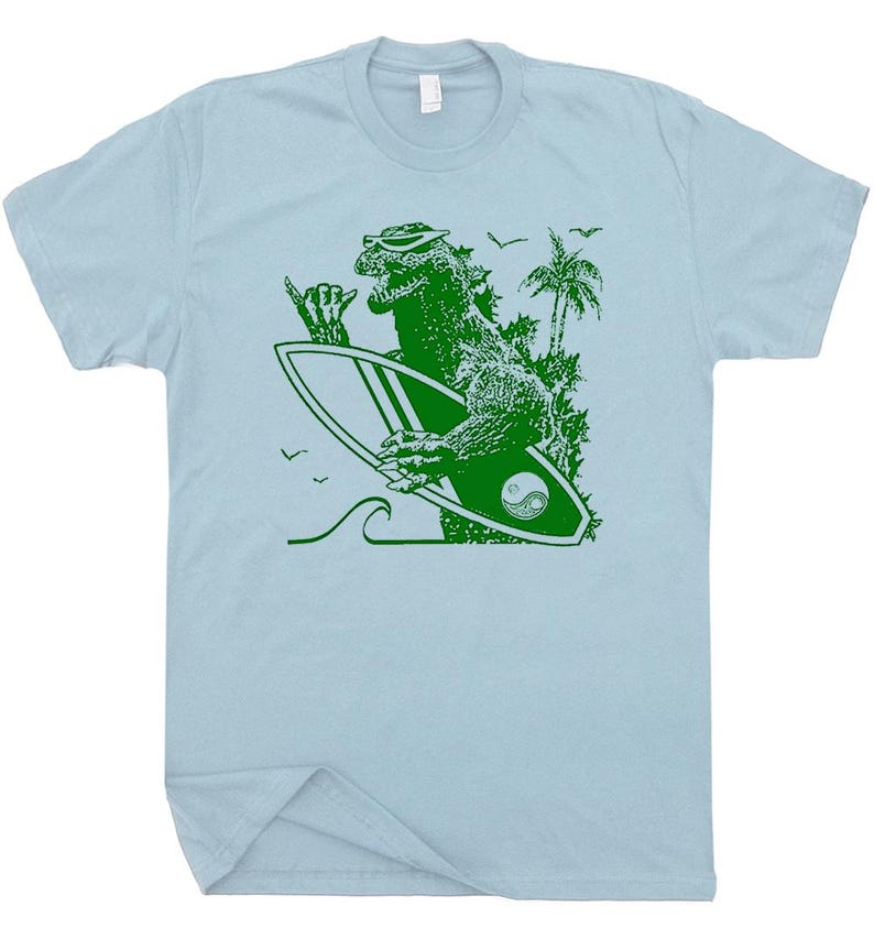 Puede incluir: Camiseta azul claro con un gr&aacute;fico verde de Godzilla con gafas de sol y una tabla de surf. La tabla de surf tiene un s&iacute;mbolo de yin-yang. Tambi&eacute;n se representan palmeras, olas y p&aacute;jaros.