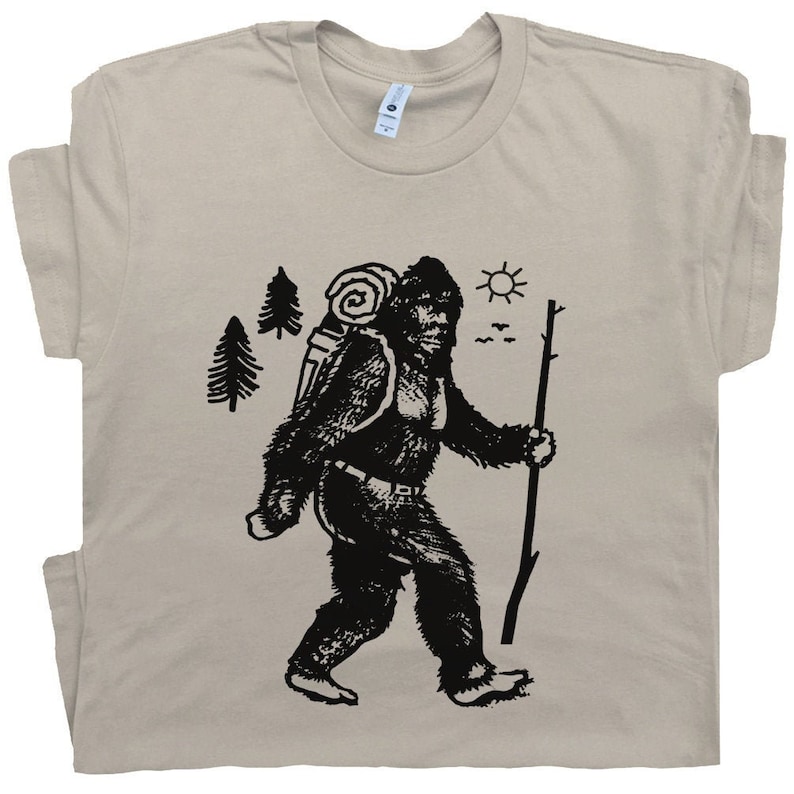 May include: A beige t-shirt with a black graphic of a Bigfoot character carrying a backpack and walking with a stick. The Bigfoot is walking to the right of the image. The graphic also includes a sun, trees, and birds in the background.
