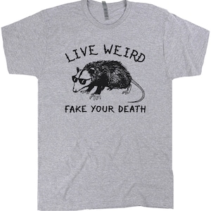 K&ouml;nnte beinhalten: Ein graues T-Shirt mit einer schwarz-wei&szlig;en Illustration eines Opossums mit Sonnenbrille. Der Text "Live Weird Fake Your Death" ist auf dem T-Shirt aufgedruckt.