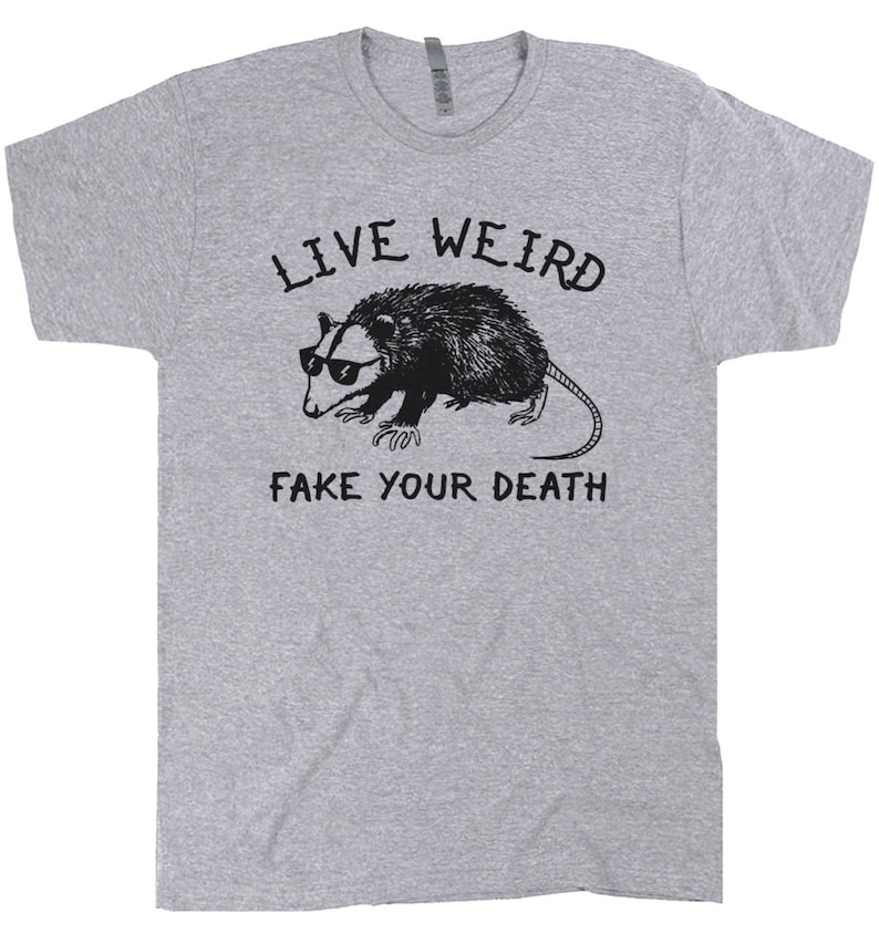 K&ouml;nnte beinhalten: Ein graues T-Shirt mit einer schwarz-wei&szlig;en Illustration eines Opossums mit Sonnenbrille. Der Text "Live Weird Fake Your Death" ist auf dem T-Shirt aufgedruckt.