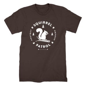 Könnte beinhalten: Ein braunes T-Shirt mit einem weißen, kreisförmigen Design. Das Design enthält die Wörter "Squirrel Patrol", "Protect" und "Defend", zusammen mit einem weißen Eichhörnchen, Bäumen und Eicheln. Der Text "Squirrel Den Studios" befindet sich darunter.