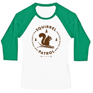 May include: A white and green baseball tee shirt with a brown graphic of a squirrel on a branch. The graphic is surrounded by a circle with the text "Squirrel Patrol" and the words "Protect" and "Defend" on either side of the squirrel.