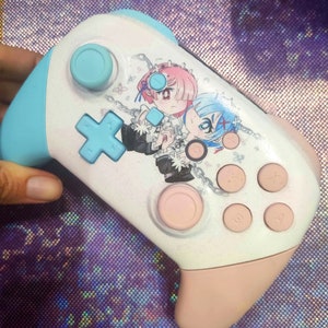 May include: A white and pink Nintendo Switch controller with a custom design featuring two anime characters. The characters are a girl with pink hair and a boy with blue hair. The controller has a blue and white button layout.