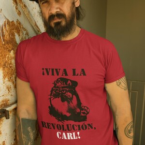 May include: A red short-sleeved t-shirt featuring a black graphic print of a cat wearing a military-style cap and glasses. The text above the cat reads ¡VIVA LA, and the text below reads REVOLUCIÓN, CARL! in a bold, dark, sans-serif font.