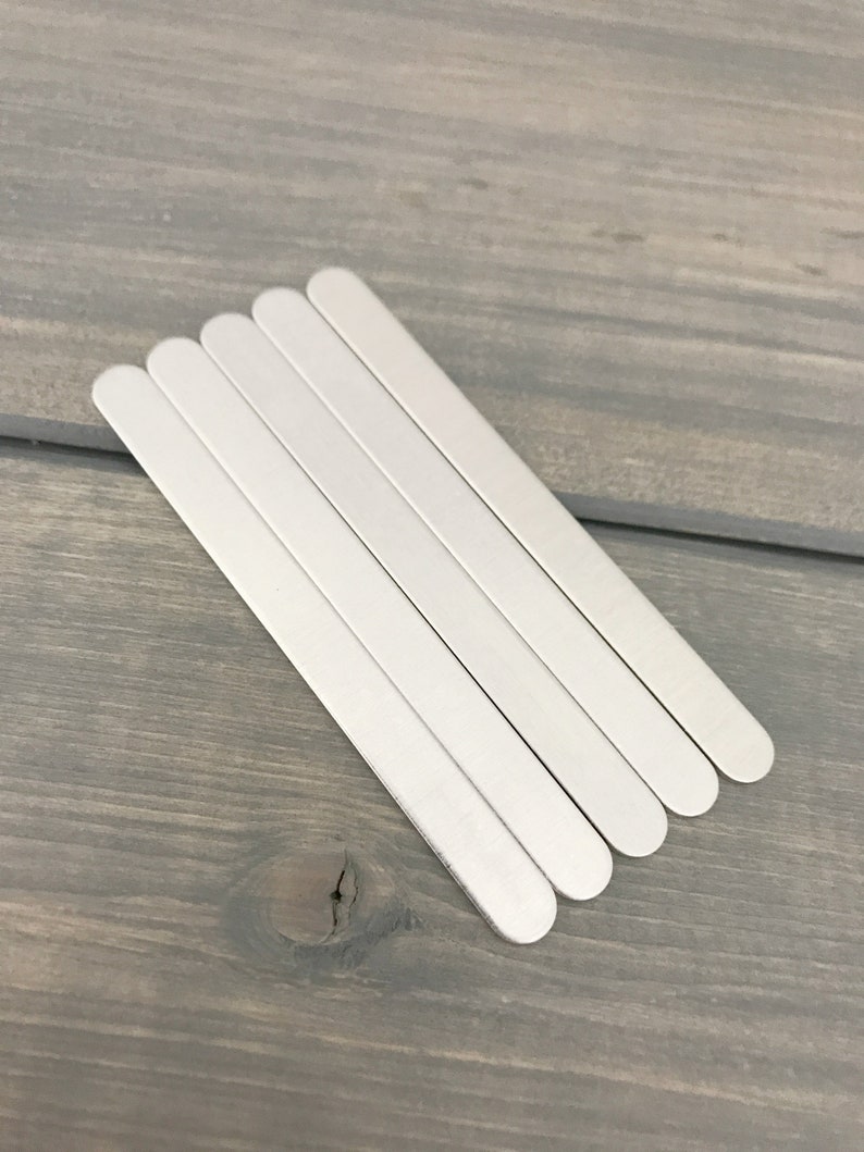 May include: Five silver metal strips, each with rounded ends, are stacked on top of each other on a gray wooden surface.