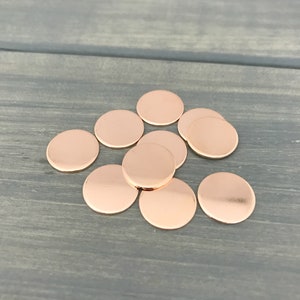 May include: A group of ten round, copper-colored metal discs. The discs are arranged in a loose pile on a wood surface.