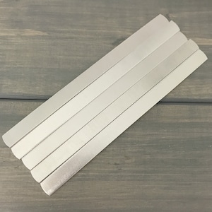 Puede incluir: Cinco tiras de metal plateado apiladas una encima de la otra. Las tiras son planas y rectangulares con bordes redondeados.