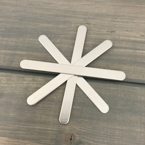 May include: A set of six silver metal craft sticks arranged in a star shape on a woodgrain surface.