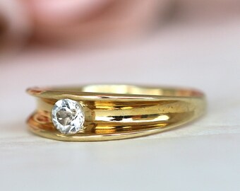 May include: A gold ring with a single, clear, round gemstone set in the center. The ring has a simple, elegant design.