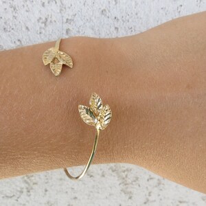 May include: A gold leaf bracelet with a delicate design. The bracelet is open and has two leaf-shaped charms.