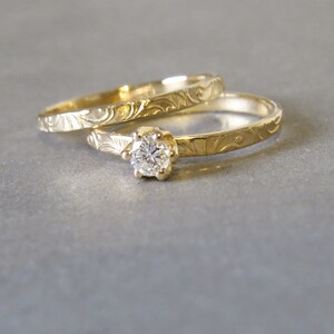 K&ouml;nnte beinhalten: Zwei goldene Eheringe mit filigranen floralen Gravuren. Der obere Ring hat einen einzelnen runden Diamanten.