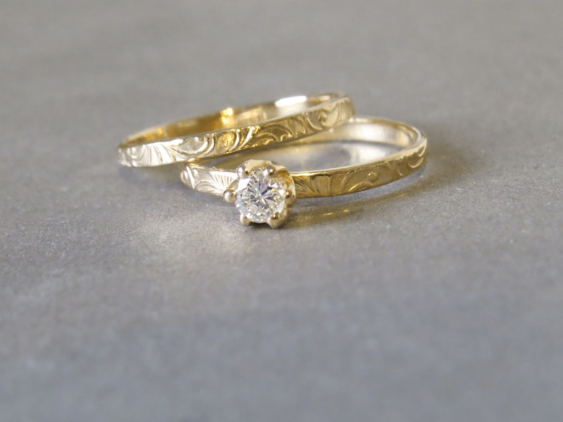 K&ouml;nnte beinhalten: Zwei goldene Eheringe mit filigranen floralen Gravuren. Der obere Ring hat einen einzelnen runden Diamanten.
