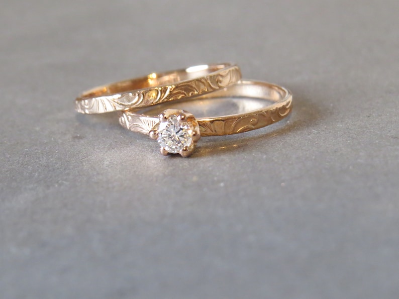 K&ouml;nnte beinhalten: Zwei goldene Eheringe mit aufwendigen Gravuren. Der obere Ring hat einen einzelnen runden Diamanten.