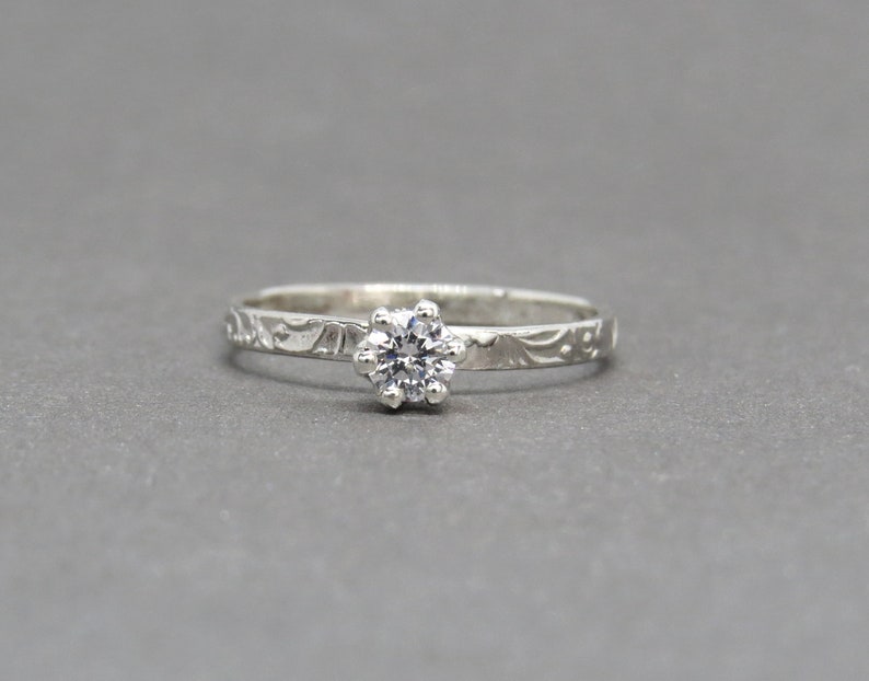 K&ouml;nnte beinhalten: Ein silberner Ring mit einem kleinen, klaren Edelstein, der in der Mitte eingesetzt ist. Die Band ist mit einem zarten, gravierten Muster verziert.