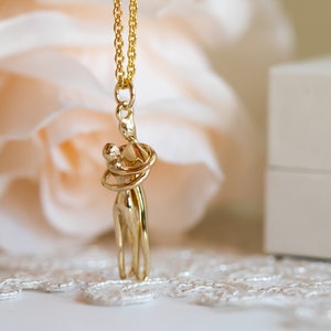 May include: Gold necklace featuring a pendant of two figures embracing. The figures are stylised and abstract, with the woman's arms wrapped around the man's neck.