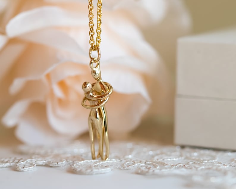 May include: Gold necklace featuring a pendant of two figures embracing. The figures are stylised and abstract, with the woman's arms wrapped around the man's neck.