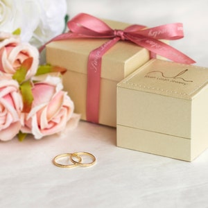 May include: Two gold wedding bands on a white surface next to a beige gift box with a pink ribbon and a bouquet of pink and white roses. The gift box has the text "Swan Lovin Jewelry" on the top.