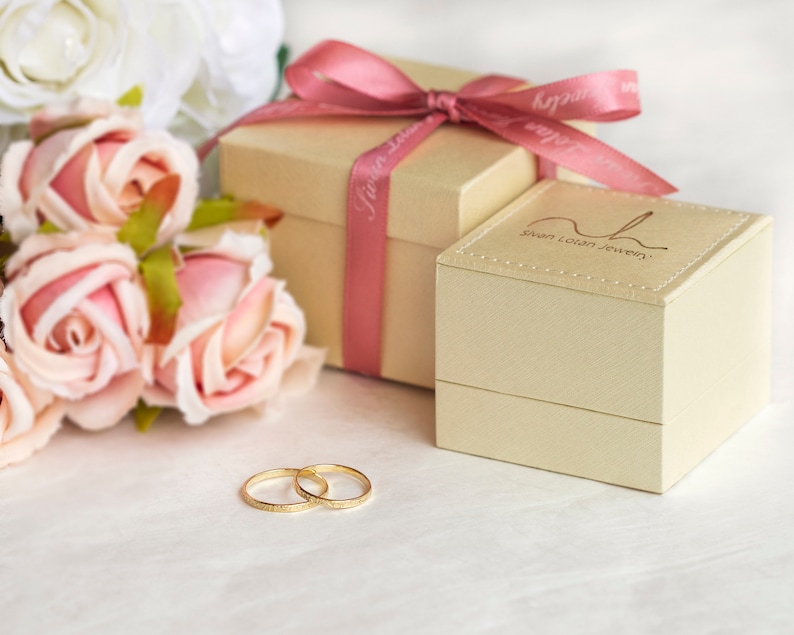 May include: Two gold wedding bands on a white surface next to a beige gift box with a pink ribbon and a bouquet of pink and white roses. The gift box has the text "Swan Lovin Jewelry" on the top.