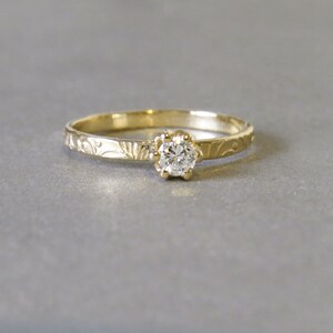 K&ouml;nnte beinhalten: Ein zarter Goldring mit einem kleinen, runden Diamanten, der in einem floralen Design gefasst ist.