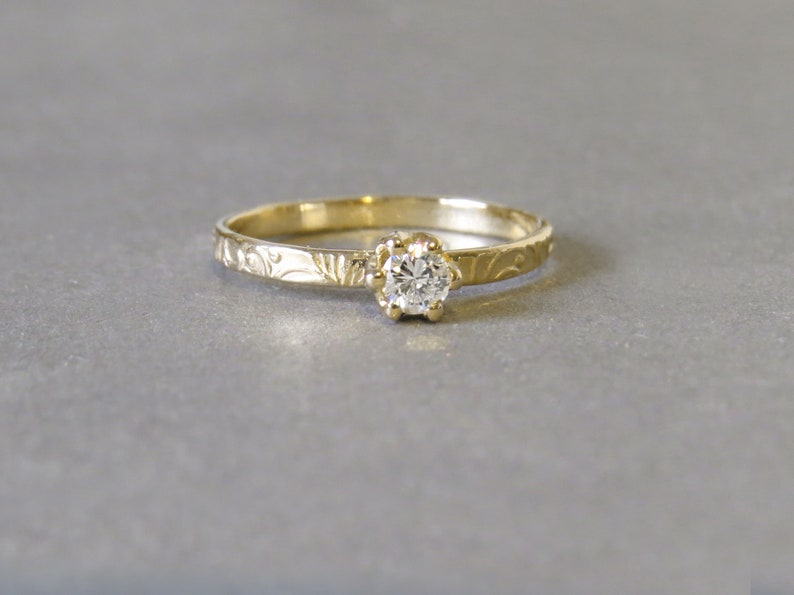 K&ouml;nnte beinhalten: Ein zarter Goldring mit einem kleinen, runden Diamanten, der in einem floralen Design gefasst ist.