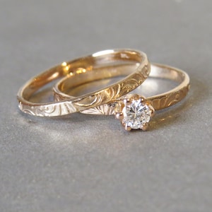K&ouml;nnte beinhalten: Zwei goldene Eheringe mit aufwendigen Gravuren. Der obere Ring hat einen einzelnen runden Diamanten.