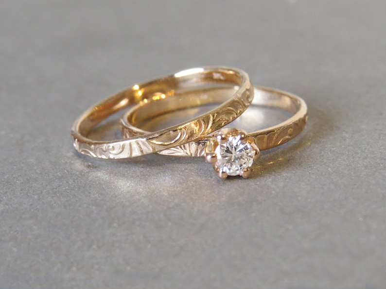 K&ouml;nnte beinhalten: Zwei goldene Eheringe mit aufwendigen Gravuren. Der obere Ring hat einen einzelnen runden Diamanten.