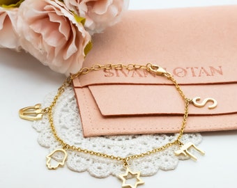 May include: A gold chain bracelet with charms including a Star of David, a hamsa hand, a Hebrew letter, and a small letter 'S'. The bracelet is on a white lace doily and a pink pouch with the text 'Sivan Otan' is in the background.