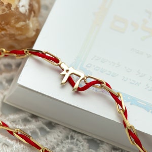 May include: A red string bracelet with a gold Hebrew letter charm. The charm is shaped like the letter 'Chai' and has a small star in the center.