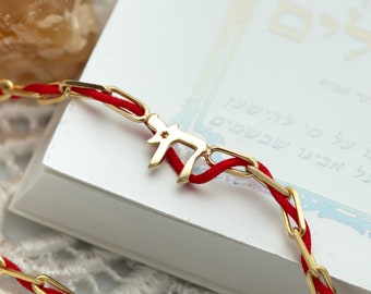 May include: A red string bracelet with a gold Hebrew letter charm. The charm is shaped like the letter 'Chai' and has a small star in the center.