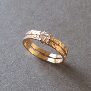 K&ouml;nnte beinhalten: Ein Paar goldene Eheringe mit einem gravierten Blumenmuster. Der obere Ring hat einen einzelnen, runden Diamanten.