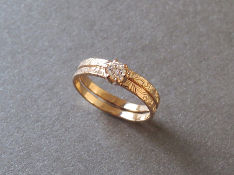 K&ouml;nnte beinhalten: Ein Paar goldene Eheringe mit einem gravierten Blumenmuster. Der obere Ring hat einen einzelnen, runden Diamanten.